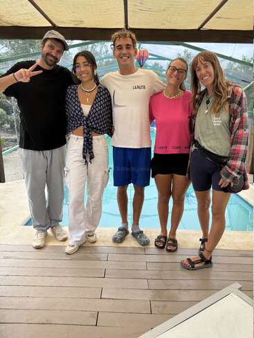 Five people are standing together, smiling by a pool under a covered patio. They are casually dressed, looking happy and relaxed, enjoying each other's company.
