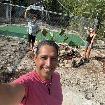 Três pessoas estão sorrindo e acenando ao ar livre. O homem na frente tira uma selfie, enquanto as outras duas ficam atrás dele, perto de um jardim de pedras, rodeadas por vegetação.