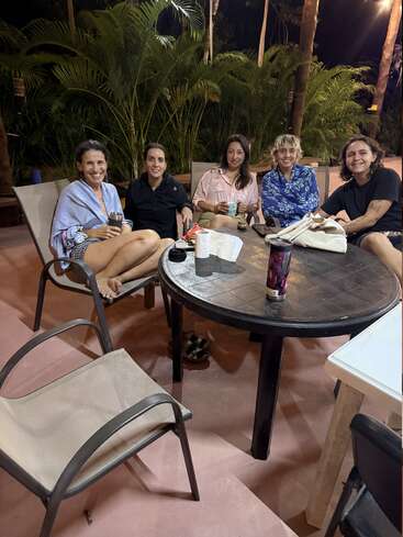Cinco pessoas estão sentadas ao redor de uma mesa redonda ao ar livre à noite, sorrindo e tomando uns drinques. Plantas e árvores verdejantes cercam o ambiente aconchegante e descontraído.