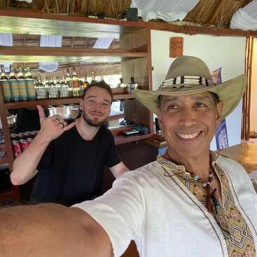 Dos hombres sonrientes en un bar; uno lleva un sombrero y una camisa bordada, el otro levanta el pulgar. Detrás de ellos se ven botellas y bebidas.