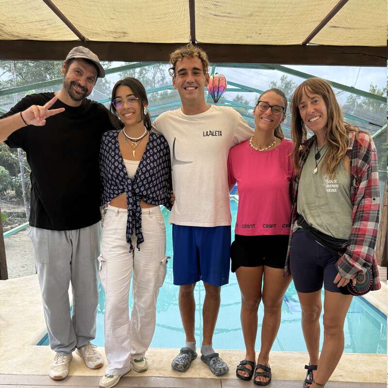 Five people are standing together, smiling by a pool under a covered patio. They are casually dressed, looking happy and relaxed, enjoying each other's company.