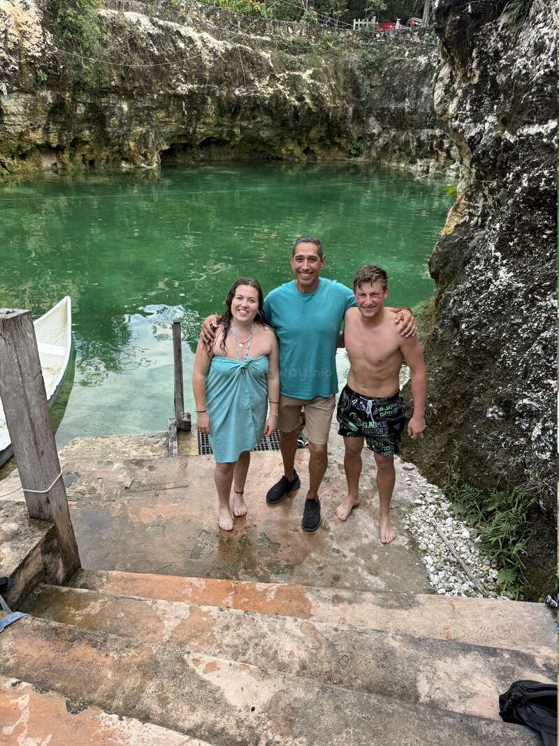 Three people, smiling and arm-in-arm, stand on stone steps by a clear green natural pool, surrounded by rocky cliffs and lush vegetation. Relaxed, tropical atmosphere.