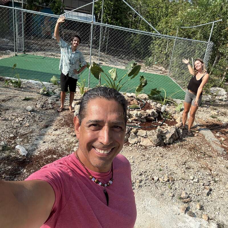 Três pessoas estão sorrindo e acenando ao ar livre. O homem na frente tira uma selfie, enquanto as outras duas ficam atrás dele, perto de um jardim de pedras, rodeadas por vegetação.