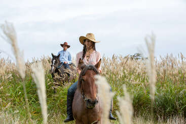 A imagem mostra duas pessoas montadas em cavalos, uma à esquerda e outra à direita, tendo como pano de fundo um campo gramado. A cena é serena e pacífica.