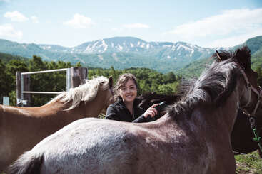 Uma pessoa está em pé na frente de um cavalo, com a cabeça do cavalo visível ao fundo. A pessoa está usando um chapéu e tem bigode. O cavalo é branco com uma crina preta.