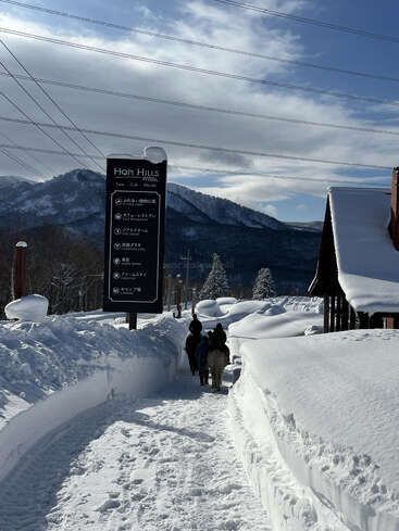 Um caminho nevado ladeado por altos bancos de neve leva às montanhas. Três pessoas a cavalo passam por baixo de fios elétricos, passando por uma placa que diz "Hopi Hills".