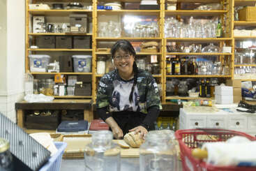 Uma mulher sorridente de óculos está em uma cozinha aconchegante, cortando pão em um balcão de madeira, cercada por prateleiras cheias de potes, garrafas e utensílios.