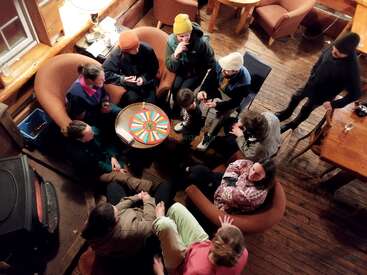 Um grupo de pessoas está reunido em uma cabana aconchegante, sentado ao redor de uma mesa jogando um jogo de tabuleiro, desfrutando de conversas e calor em um ambiente confortável.