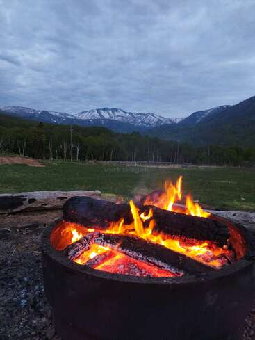 Uma fogueira ardente queima intensamente em uma fogueira de metal, cercada pela natureza. As montanhas cobertas de neve e o céu nublado criam um fundo cênico impressionante.