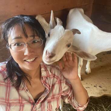 The image shows a woman with dark hair and glasses, wearing a plaid shirt, posing with a white goat in a barn or stable.