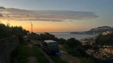 A van is parked on a grassy path overlooking a coastal town at sunset, with orange skies, hills, and a few scattered clouds in the background.