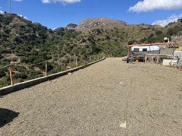 A dirt clearing overlooks green, hilly terrain under a bright blue sky. To the right, a trailer and scattered building materials sit along a simple rope fence.