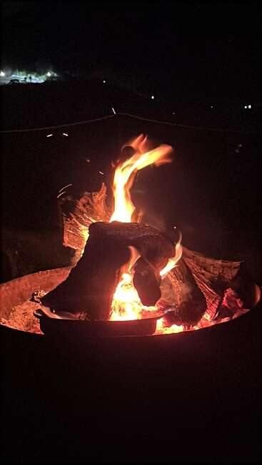 A cozy campfire burns brightly in the darkness, illuminating stacked logs. Sparks rise, and distant lights twinkle in the background, creating a peaceful, outdoor night scene.