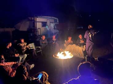 A group of people sit around a warm campfire at night, chatting and laughing, with a camper van in the background, creating a cozy outdoor ambiance.