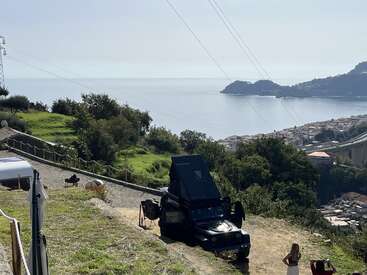 A black off-road vehicle with a rooftop tent is parked on a grassy hill overlooking the sea, with trees, a coastal town, and two people nearby.