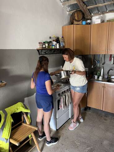 Two women are cooking together in a rustic kitchen. One is stirring food in a pot on the stove while the other assists, sharing a cozy, collaborative moment.