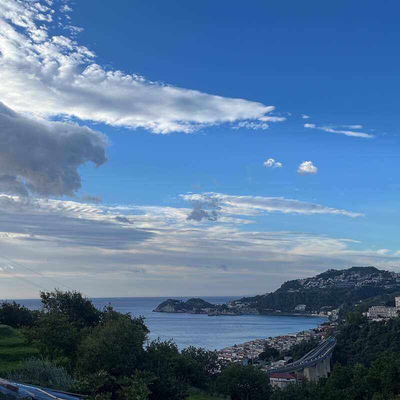 A stunning coastal view features lush green hills, a winding road, a quaint seaside town, calm blue waters, and a vibrant sky filled with dramatic clouds.
