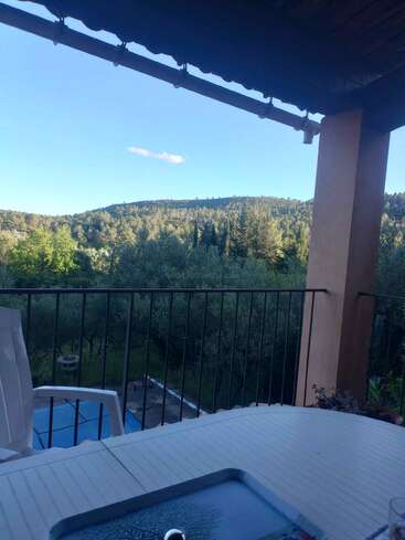 A peaceful terrace overlooks a lush green forest and distant hills, under a clear blue sky. A swimming pool, chairs, and a table are visible.