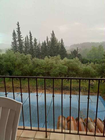 A rainy day view from a balcony shows tall pine trees, lush greenery, a blue-covered swimming pool, iron railing, and a white plastic chair in front.