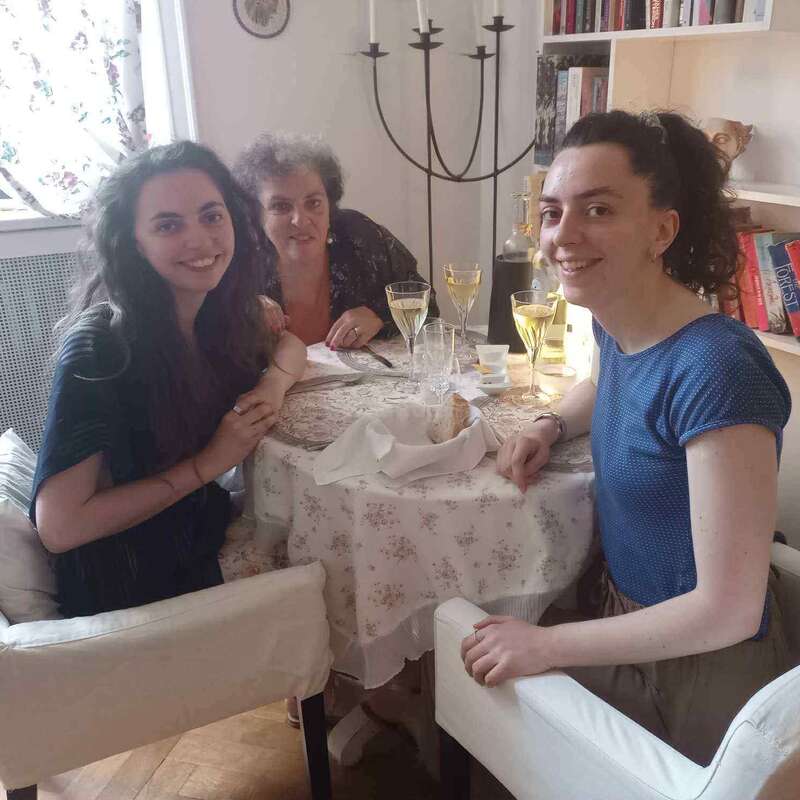 Three women sit around a round dining table with a floral tablecloth, smiling warmly. They share drinks and bread, enjoying a cozy, intimate meal together indoors.