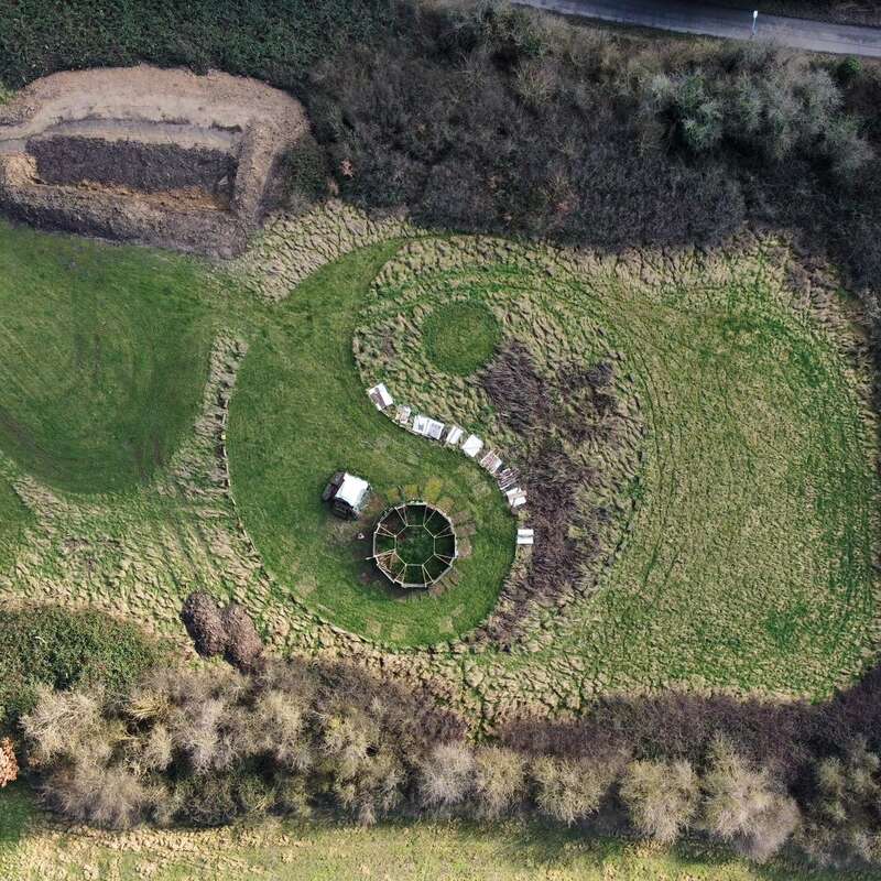 The image depicts an aerial view of a grassy field with a circular structure, surrounded by trees and bushes, featuring a winding path and a small building in the center.
