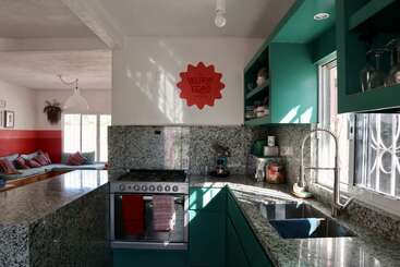 This image shows a modern kitchen with green cabinets, granite countertops, and a gas stove. Adjacent is a cozy living area with colorful cushions.