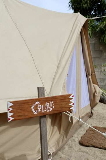 Une tente en toile beige est dressée à l'extérieur, sur le sable. Devant, un panneau en bois indique « Colibri » avec des accents décoratifs blancs et rouges. Des plantes et un mur à proximité.