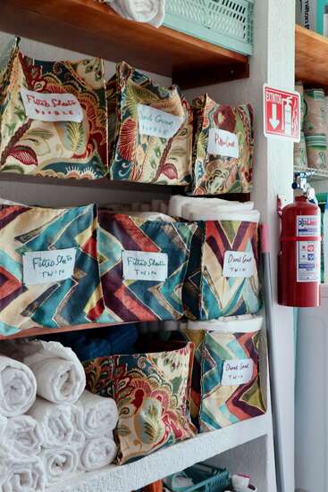 Des bacs en tissu colorés étiquetés pour le linge de maison, les draps housses et les housses de couette sont soigneusement rangés sur des étagères. Des serviettes blanches enroulées et un extincteur sont visibles.