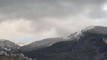 Ein dramatischer Himmel hängt über einer bergigen Landschaft mit dunkelgrünen Wäldern und Schneeflecken auf den Gipfeln unter dicken, grauen Wolken, die auf bevorstehendes Wetter hindeuten.