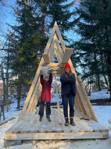 A child and an adult stand on a wooden A-frame structure in snowy woods; the child joyfully throws snow upwards while the adult smiles, arms crossed.