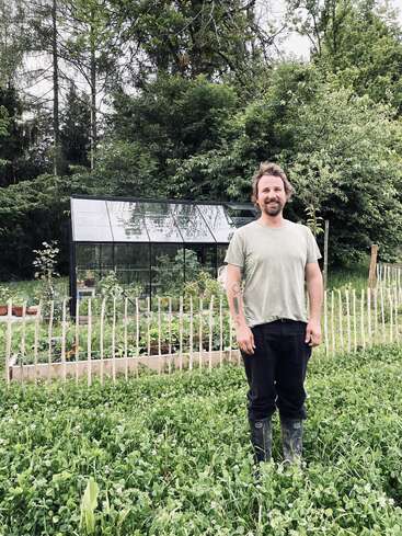 A man stands in a lush green garden with tall grass, wearing a light shirt and boots. Behind him is a greenhouse and wooden fence.
