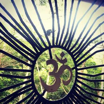 Un portail métallique avec le symbole "Om" au centre, des lignes rayonnantes comme des rayons de soleil, encadrant des arbres verdoyants et la lumière du jour en arrière-plan.