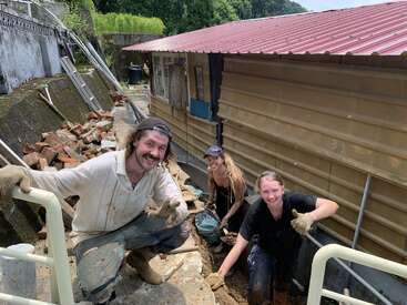 Três pessoas sorriem e fazem sinal de positivo enquanto trabalham ao ar livre em um prédio de metal bronzeado. Elas parecem enlameadas, cercadas por tijolos, ferramentas, escadas e materiais de construção.