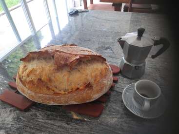 Ein frischer, knuspriger Brotlaib liegt auf einem Holzbrett neben einer Mokkakanne und einer leeren Tasse, alles auf einer Marmorarbeitsplatte in der Nähe der sonnenbeschienenen Fenster.