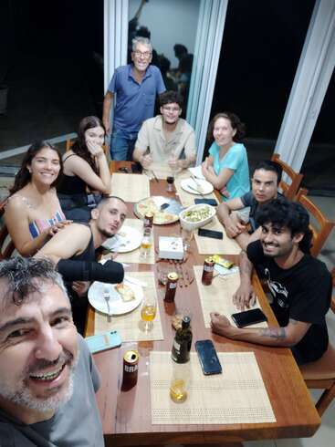 Eine Gruppe von neun Personen ist um einen Tisch versammelt, lächelt und genießt das Essen und die Getränke in einem Raum. Es scheint ein fröhliches, lebhaftes Beisammensein zu sein.