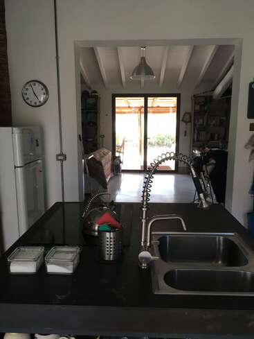 Esta imagem mostra uma cozinha moderna com pia dupla, bancada preta, torneiras de metal, utensílios e prateleiras. Mais adiante, há uma sala de estar e portas de vidro.