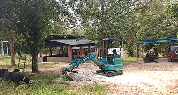 Ein kleiner grüner Bagger arbeitet in der Nähe einer unbefestigten Straße, umgeben von Bäumen und Grünpflanzen. Im Hintergrund sind Backsteingebäude, geparkte Fahrzeuge und noch mehr Vegetation zu sehen.