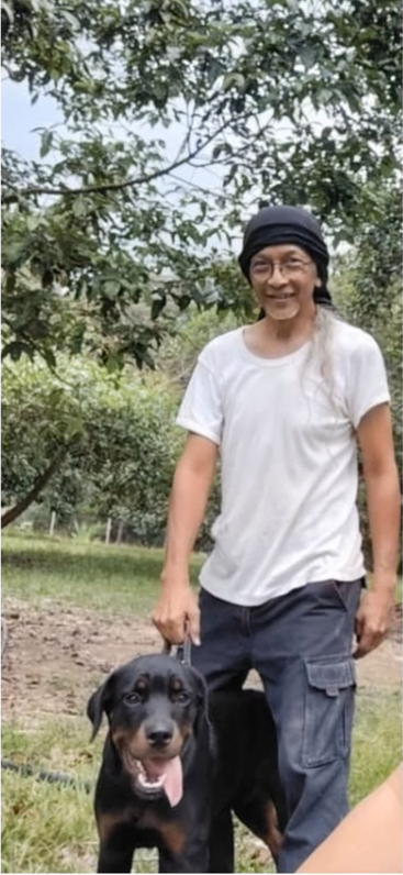 Ein lächelnder Mann mit Brille und schwarzem Kopftuch steht im Freien und hält einen glücklichen schwarz-braunen Hund an der Leine, umgeben von grünen Bäumen.