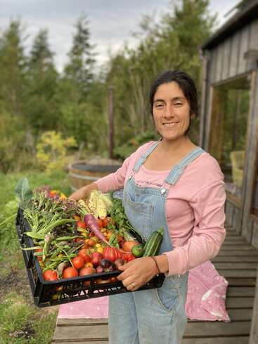 Uma mulher sorridente, vestindo macacão, está do lado de fora segurando uma grande caixa cheia de vegetais recém-colhidos, incluindo tomates, feijões, abobrinhas e folhas verdes. Uma vegetação exuberante a rodeia.