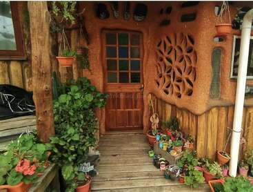 Este acogedor porche rústico cuenta con una puerta de madera, paredes esculpidas en tierra, plantas en macetas, decoración única, macetas colgantes y vigas de madera natural que crean una entrada artística y acogedora.