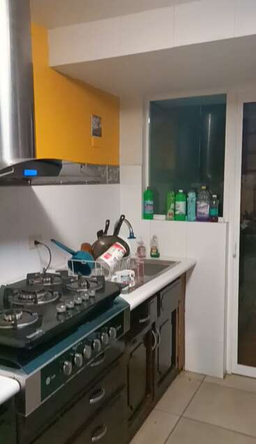 Das ist eine Küche mit einem schwarzen Herd, einem gelben Schrank, einer Spüle und verschiedenen Reinigungsflaschen am Fenster. Auf dem Abtropfgestell trocknet das Geschirr.