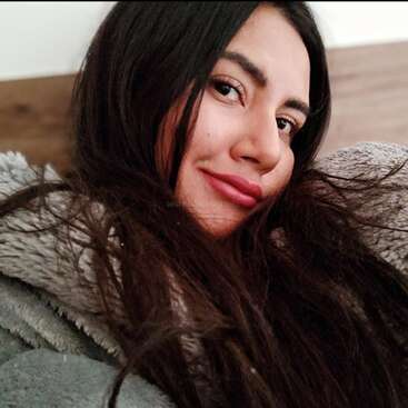 Une jeune femme aux longs cheveux bruns est allongée, vêtue d'une veste grise confortable, et sourit doucement, avec une tête de lit en bois et un mur blanc en arrière-plan.