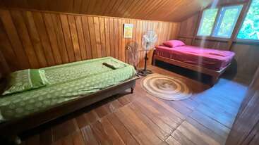 Este aconchegante quarto de madeira tem duas camas de solteiro com roupa de cama verde e vermelha, um ventilador de pé, um tapete redondo, um quadro emoldurado, janelas e uma luz natural acolhedora.