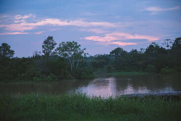 Une scène fluviale sereine au crépuscule, entourée d'arbres et d'herbes verdoyants, sous un ciel éclatant aux nuances de bleu, de violet et de rose.