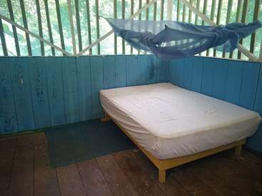 Une chambre simple et rustique aux murs en bois bleu, avec un lit sobre recouvert d'un drap blanc, une moustiquaire au-dessus, et la lumière du soleil qui pénètre à travers les fenêtres à persiennes, créant une atmosphère paisible.