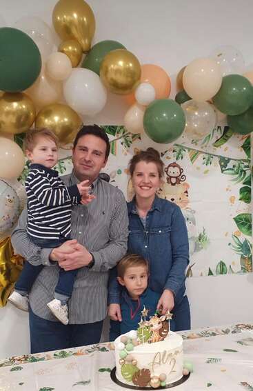 Une famille de quatre personnes pose en souriant lors d'une fête d'anniversaire. On voit des ballons, des décorations sur le thème de la jungle et un gâteau d'anniversaire sur lequel est inscrit « Alan 2 » sur la table.