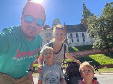 Une famille de quatre personnes profite d'une journée ensoleillée pour faire du tourisme, avec deux enfants, une poussette et, en arrière-plan, un bâtiment historique orné de tours et entouré de verdure.