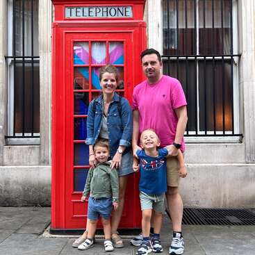 Una familia de cuatro miembros, todos sonrientes, posa delante de una clásica cabina telefónica roja británica en una calle de la ciudad. Los padres y los hijos parecen felices y relajados.