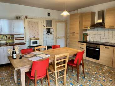 In dieser gemütlichen Küche gibt es einen Holztisch mit gemischten Stühlen, rote und hölzerne, alte Fliesen, moderne Geräte, weiße Schränke, eine Weinflasche und Papierkram.