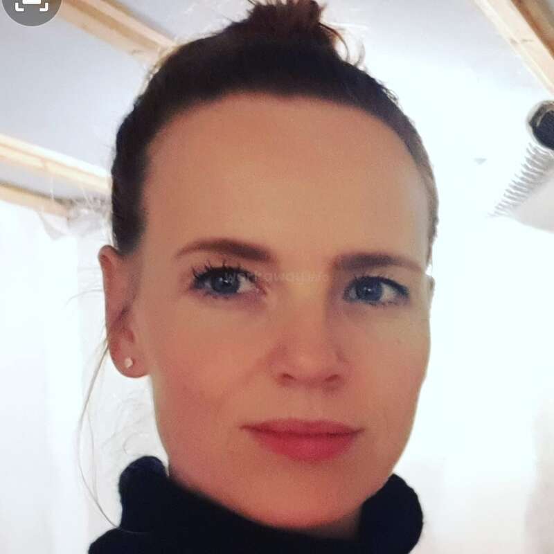 Eine Frau mit blauen Augen und heller Haut schaut direkt in die Kamera. Sie hat ihr Haar zu einem Dutt gebunden und trägt ein schwarzes Oberteil.